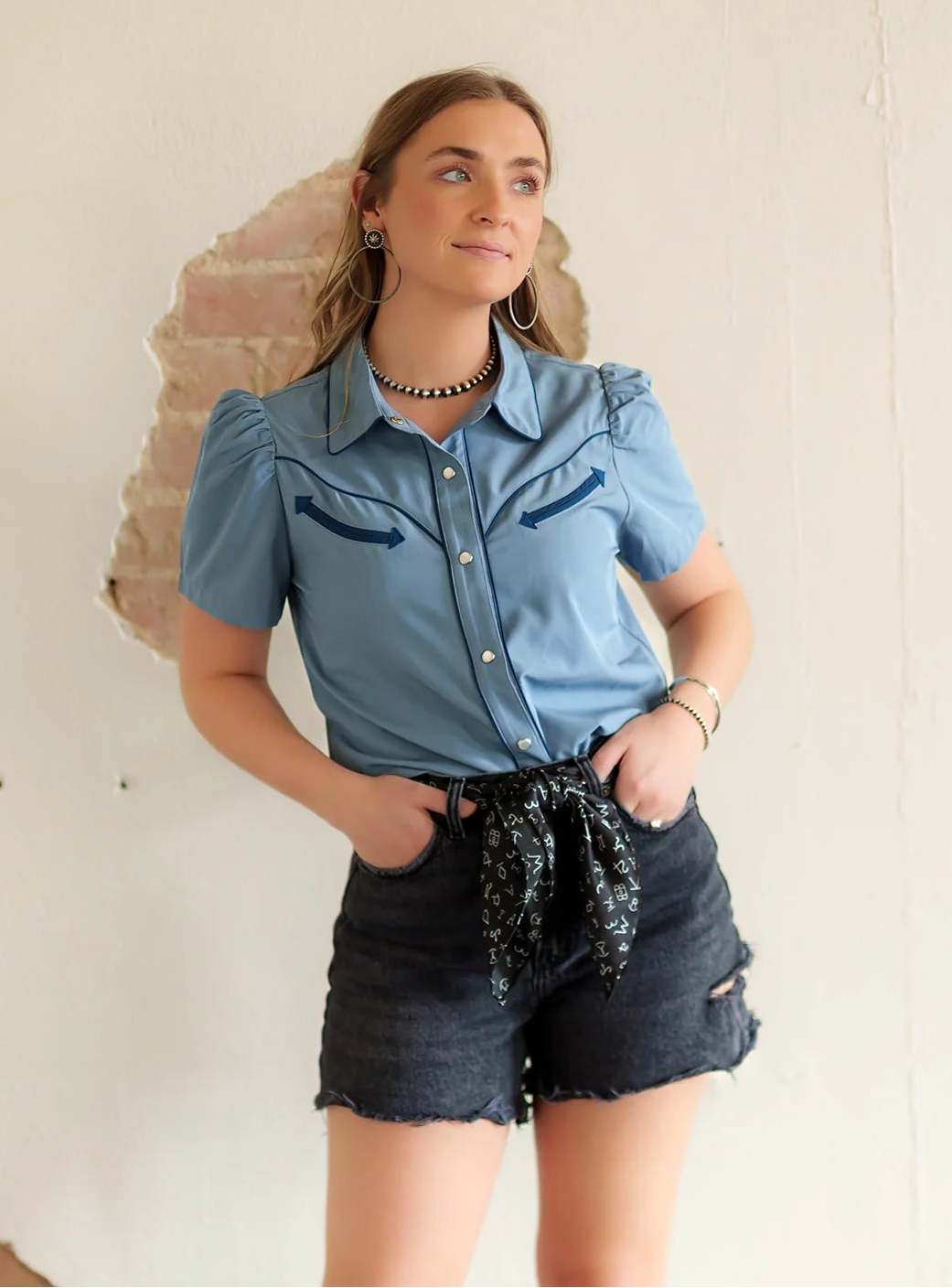 Woman wearing a blue shirt and denim shorts against a textured wall.