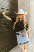 Woman wearing a cowboy hat and black top with a denim skirt, standing against a wall.